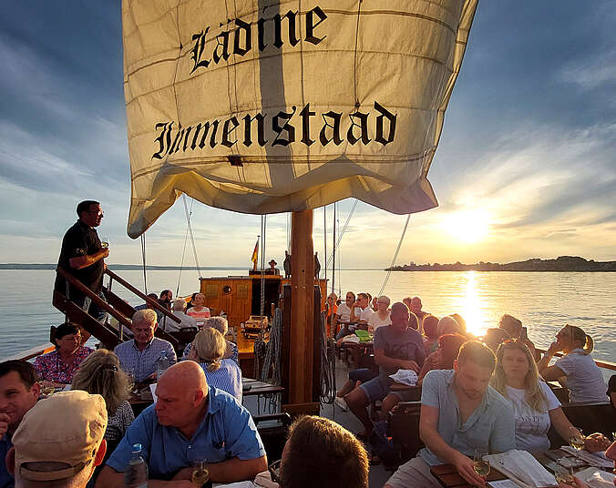 Frauen und Männer sitzen auf einem Segelboot an Tischen bei einer Weinprobe.