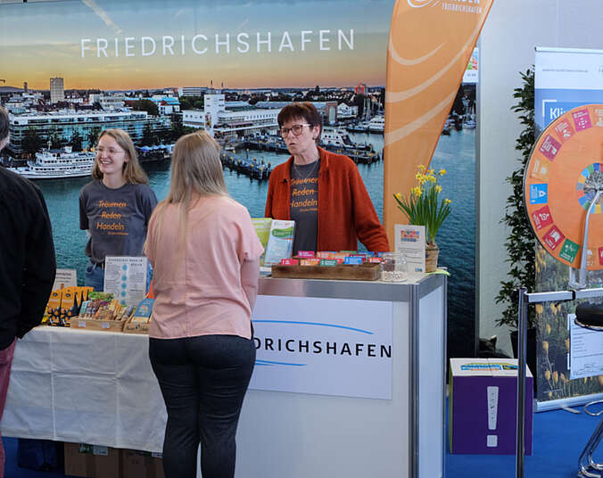 Zwei Frau stehen hinter einer Verkaufstheke im Gespräch mit Besuchern. Im Hintergrund eine Foto von Friedrichshafen.