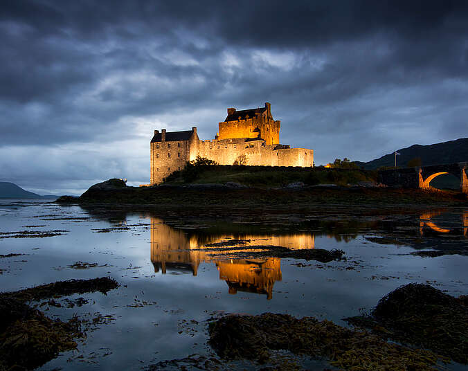 Eine in der Abenddämmerung hellbeleuchtete rustikale Burg auf einer kleinen Insel in einer Meeresbucht. Es führt eine steinerne Brücke vom Land zur Insel, alles ist umgeben von Wasser.