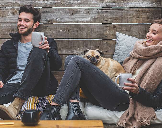 Zwei junge Leute sitzen warm eingepackt mit einer Tasse auf einem Outdoor-Sofa.