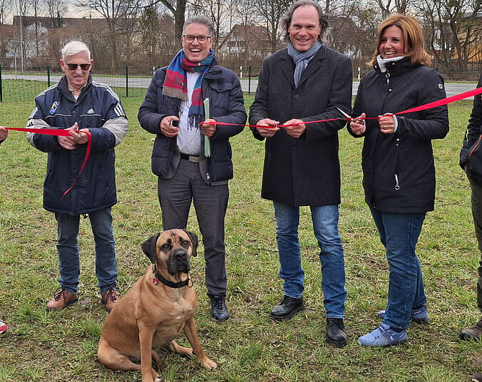 Drei Männer und eine Frau durchtrennen ein rotes Band. Im Vordergrund sitzt ein Hund.