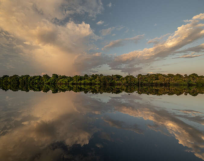 Himmel, Wolken und Uferzone mit Bewaldung spiegeln sich in einer galtten Wasseroberfläche eins zu eins.