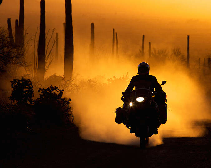 Ein Mann auf einem Motorad fährt in einer Staubwolke auf einer trostlosen Landstraße im Nirgendwo. Man erkennt nur Silhouetten.